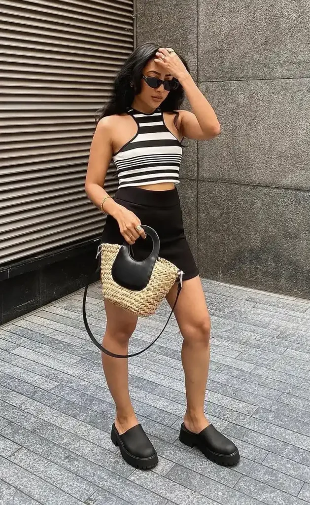 Petite woman in a black and white horizontal stripe halter racerback crop top and a black bodycon mini skirt, holding a round woven straw bag with a black leather arc handle, wearing black chunky lug sole platform mules and small dark oval sunglasses, with stacked gold rings and a gold bracelet, posing against a stone wall and corrugated metal backdrop.