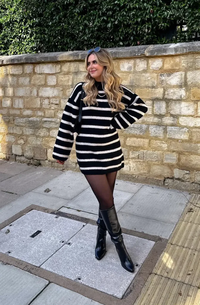 Petite blonde woman in a bold black and white horizontal stripe oversized mini sweater dress with a crew neckline and long sleeves, paired with black sheer tights, tall sleek black knee-high boots with a slender heel, and small dark sunglasses perched on her head, standing on a stone paved sidewalk in front of a golden limestone garden wall with dense green hedging overhead.