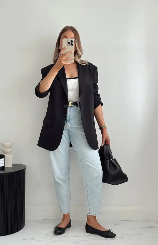 Woman taking a mirror selfie in very pale bleached high-waisted ankle-cropped mom jeans, a white ribbed square-neck fitted top with a black trim neckline, an oversized black blazer worn open with sleeves pushed to the elbow, a black belt with a gold logo-style buckle, black bow-detail ballet flats, and a soft black drawstring bucket bag, in a minimal white interior
