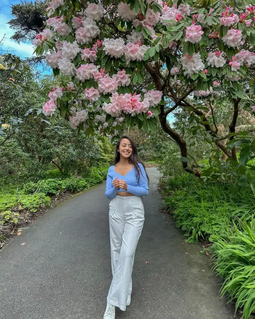 Petite woman walking on a garden path under a blossoming pink rhododendron tree, wearing a periwinkle-blue fitted long-sleeve crop top with a sweetheart neckline and high-waisted white wide-leg trousers with white boots, greenery on both sides.