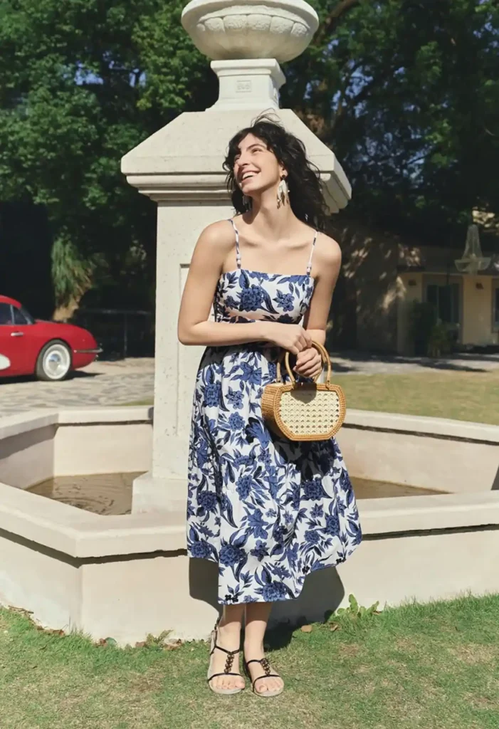 Petite woman in a white and navy blue large floral spaghetti strap midi dress with a full skirt, holding a small structured woven rattan bag with bamboo handles, wearing strappy dark sandals and chandelier drop earrings, standing near a stone fountain in a bright sunny outdoor setting with green trees in the background.