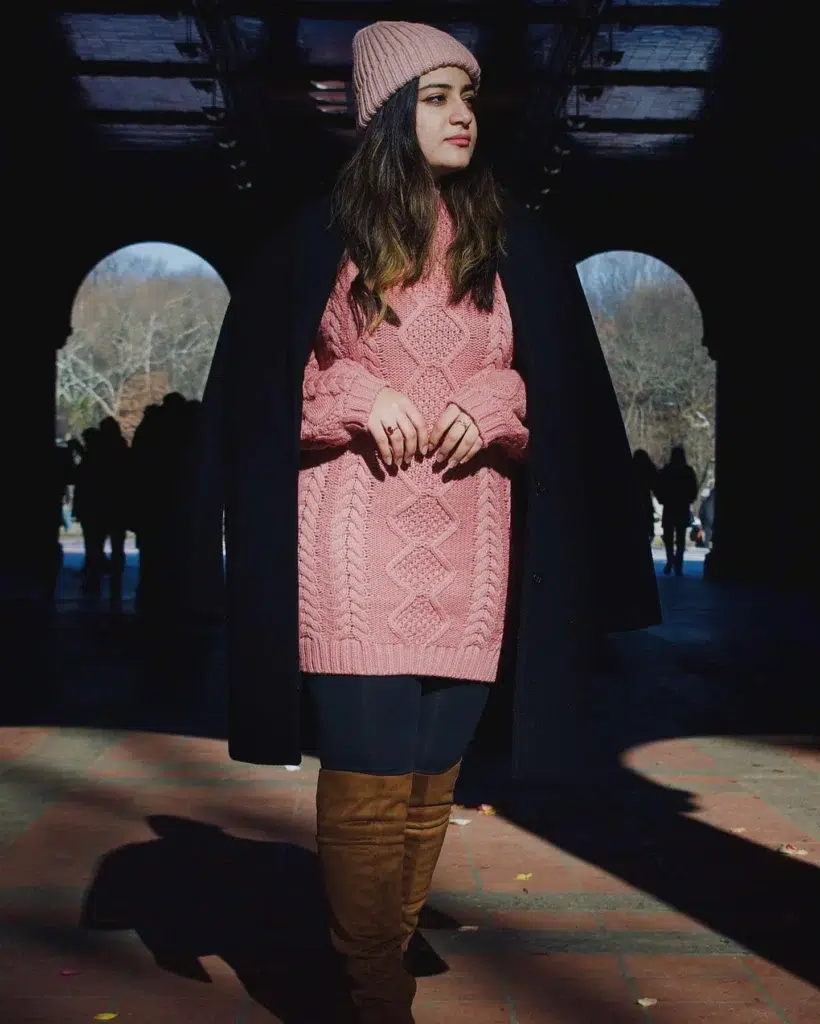 Petite pear-shaped woman in a chunky blush pink cable-knit tunic sweater, dark fitted leggings, and tan suede over-the-knee boots, wearing a dark navy wool coat draped open over her shoulders and a matching pink ribbed beanie, standing in a sunlit stone archway in a park.