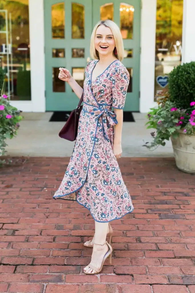 Petite woman in a blush pink floral wrap midi dress with blue contrast piping trim outlining the deep V-neckline, wrap front, and asymmetric handkerchief hem, with elbow-length sleeves and a self-tie waist bow, wearing nude strappy stiletto sandals and carrying a small structured burgundy top-handle bag, with dangling dark red earrings, standing on red brick pavers in front of a boutique with sage green painted doors and large potted floral arrangements on either side.