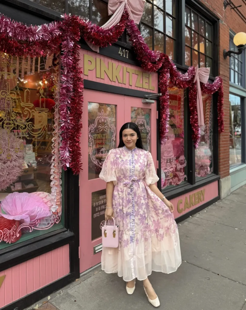A petite woman wearing a blush and cream organza floral maxi dress with purple floral print, a mandarin collar, small button-front placket, elbow-length balloon sleeves with cinched cuffs, and a self-tie fabric belt at the waist, styled with a small structured blush pink top-handle bag with gold hardware and cream square-toe low block heel pumps, photographed in front of a pink decorated bakery storefront.