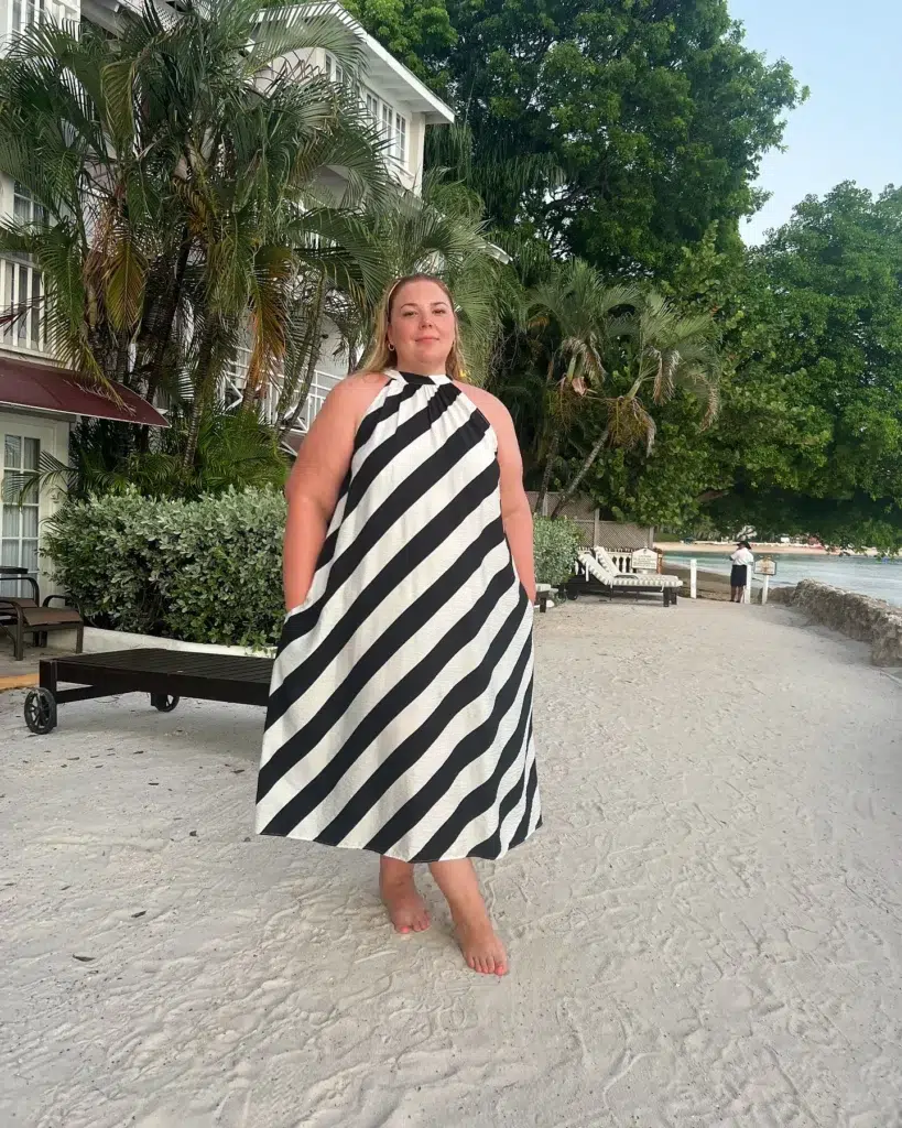 Petite plus-size woman with blonde hair worn down standing barefoot on white resort beach sand wearing a loose halter-neck A-line maxi dress in bold black and white wide diagonal stripes with a gathered twisted knot at the halter neckline and side pockets, with small gold stud earrings, set against a tropical backdrop of palm trees, a white colonial-style resort hotel building, and beach loungers with a glimpse of calm ocean water in the distance.
