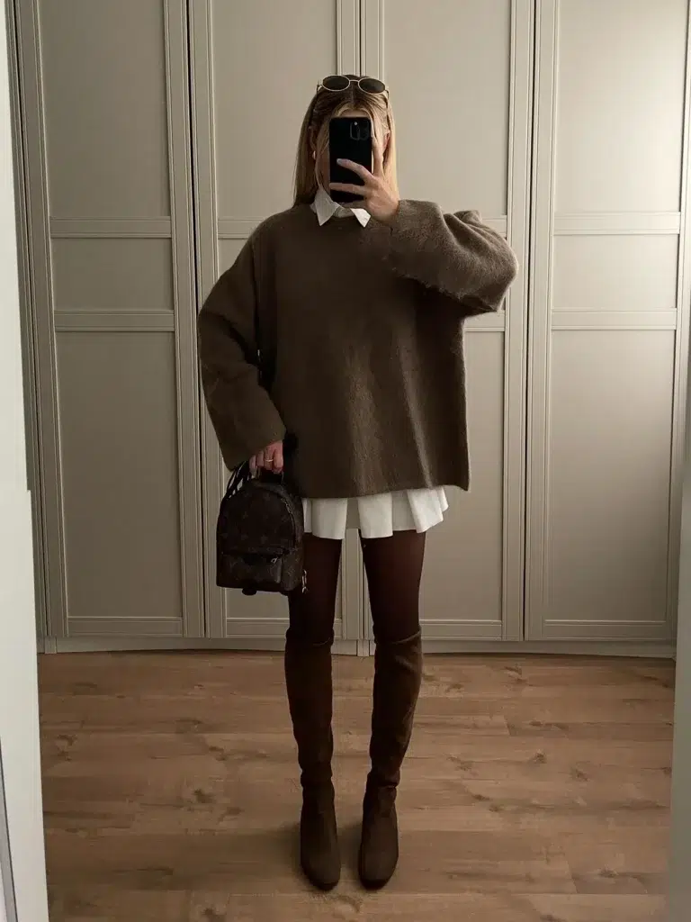 Petite woman in a boxy oversized mocha-brown brushed knit sweater worn over a white shirt dress, with a crisp white collar visible at the neckline and a strip of white hem showing below the sweater, paired with dark brown opaque tights and over-the-knee dark brown suede boots, holding a small dark monogram mini backpack, taking a mirror selfie in front of a paneled wardrobe with wooden floors.