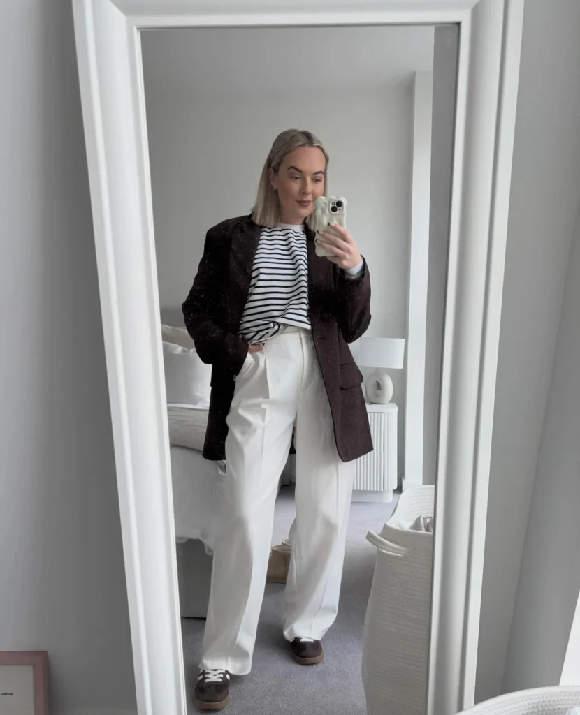 Petite woman taking a mirror selfie in a bright bedroom, wearing a navy Breton stripe tee tucked into high-waisted white wide-leg trousers, an open flecked brown blazer, and brown suede sneakers, with a bed and fluted nightstand behind her.