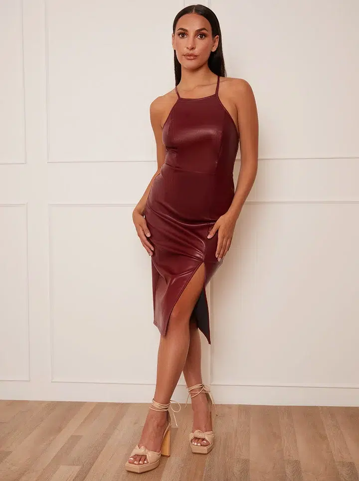 Slim brunette woman with sleek dark hair pulled back low wearing a deep burgundy faux leather halter bodycon midi dress with thin cross-back straps, a smooth fitted bodice, and an asymmetric side split hem that rises higher on one side and falls to midi length on the other, paired with cream chunky open-toe platform heels with ribbon lace-up ankle ties, photographed against a clean white paneled interior wall with light wood flooring.