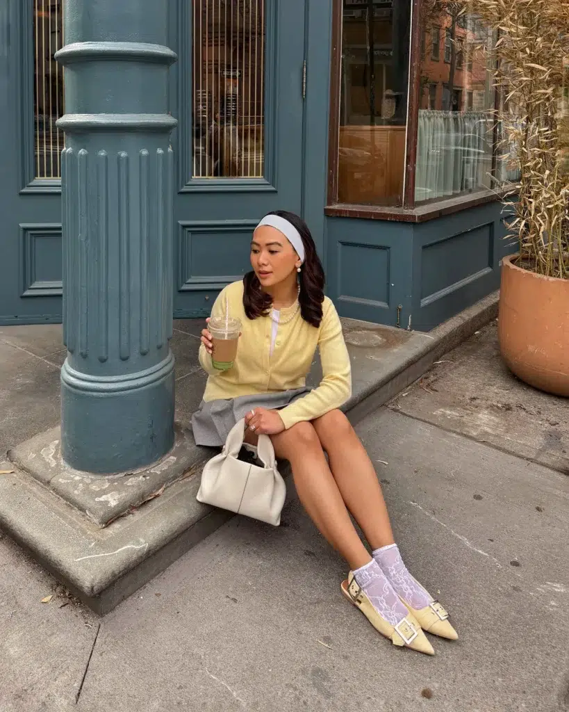 Petite woman sitting on a city curb, wearing a butter-yellow buttoned cardigan, a grey pleated mini skirt, a white headband, and a pearl necklace, holding an iced coffee, with a cream top-handle bag beside her and lavender lace socks worn over pointed buckled flats.