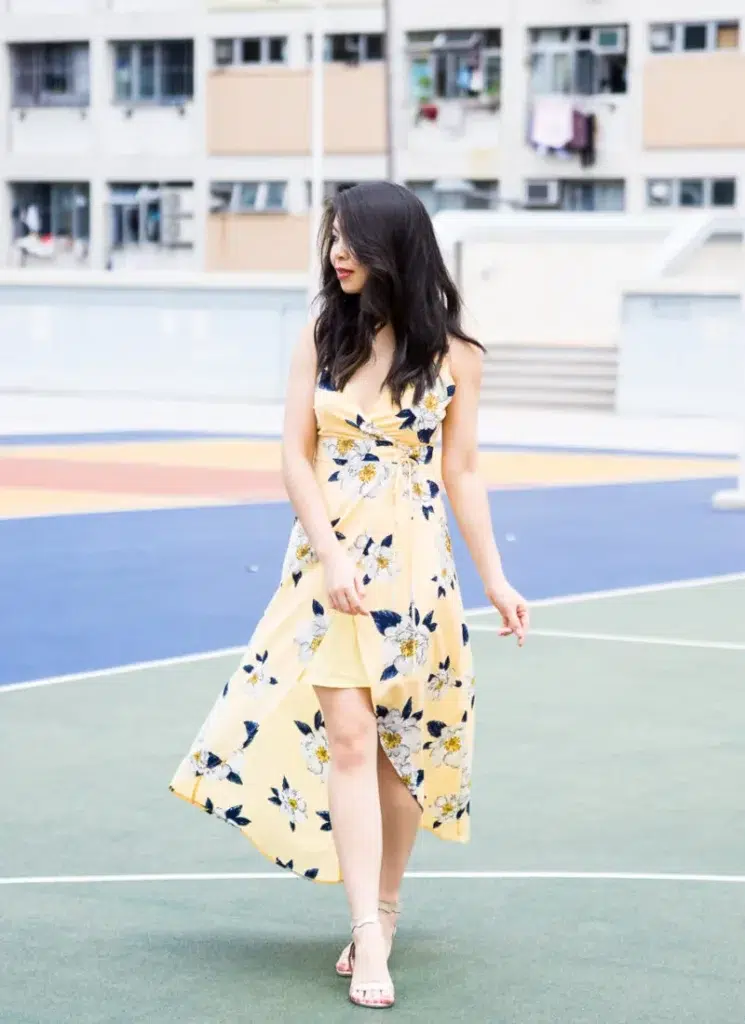 Petite woman with long dark wavy hair in a butter yellow high-low floral dress featuring large white blooms with navy leaves, a V-neckline, thin straps, and a cinched tie detail at the front waist, wearing gold strappy heeled sandals, captured mid-movement on a colorful outdoor sports court with an urban apartment building in the background.
