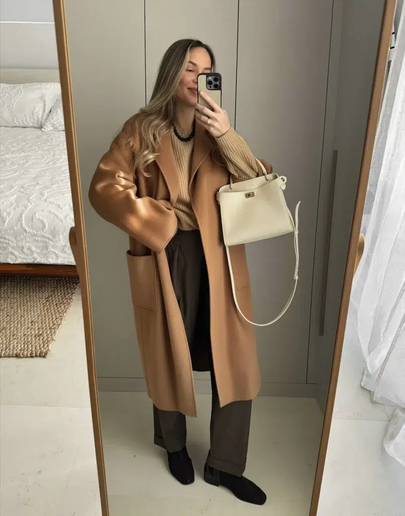 Petite woman in an oversized longline camel wool coat worn open over a camel ribbed knit sweater, paired with deep olive-brown wide-leg cuffed trousers and flat black loafers, carrying a cream structured leather top-handle bag with gold hardware and a long shoulder strap, wearing a black beaded necklace, taking a mirror selfie in a minimalist bedroom with gray wardrobe doors and a gold-framed full-length mirror.