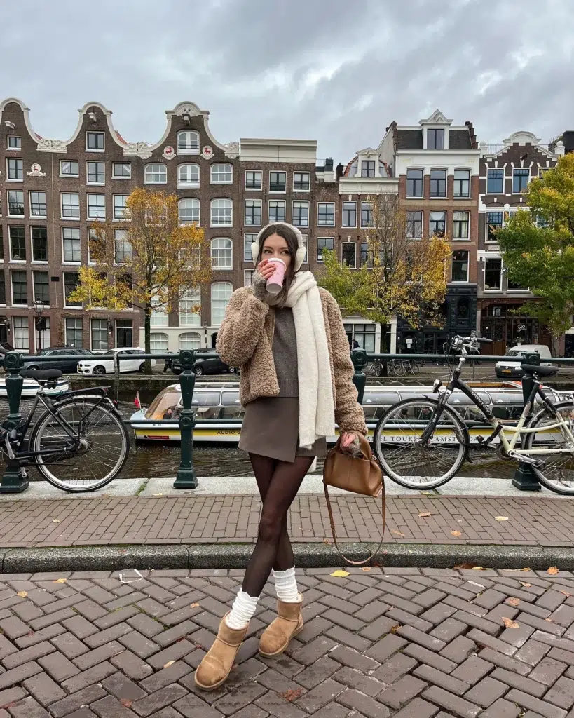Petite woman standing on a cobblestone bridge in Amsterdam with Dutch canal houses, autumn trees, and bicycles in the background, wearing a cropped camel faux-shearling teddy jacket worn open over a grey knit top and mocha brown mini skirt, dark brown opaque tights, white ribbed crew socks scrunched at the ankle above short camel UGG-style boots, cream fuzzy earmuffs, a large cream oversized scarf draped over the jacket, carrying a small structured tan leather mini bag and holding a pink takeaway coffee cup.
