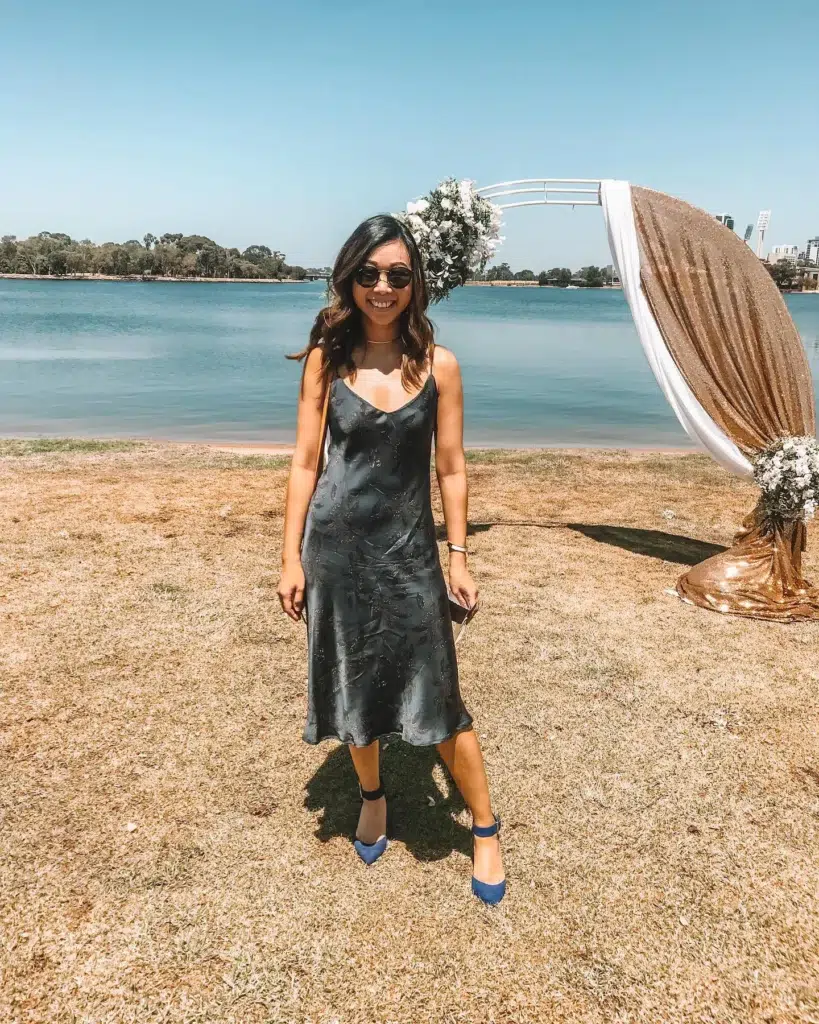 Petite Asian woman with dark wavy hair smiling outdoors at a waterfront wedding venue, wearing a charcoal grey satin midi slip dress with a subtle dark burnout floral pattern and thin spaghetti straps, paired with cobalt blue pointed-toe ankle-strap heels, a delicate gold choker necklace, small round dark sunglasses, and holding a small clutch, standing on dry grass in front of a circular wedding arch decorated with white flowers and gold sequin fabric with a blue lake and sky behind her.