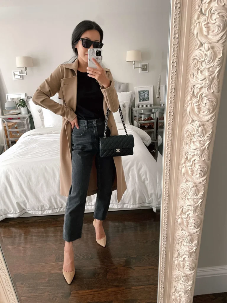 Petite woman taking a mirror selfie in a bedroom wearing high-waisted charcoal grey straight leg jeans with a raw cropped hem, a fitted black long-sleeve top, an open camel trench coat, nude pointed-toe pumps, and a black quilted crossbody bag with a silver chain strap and silver hardware, with oversized black sunglasses and hair pulled back in a low updo.