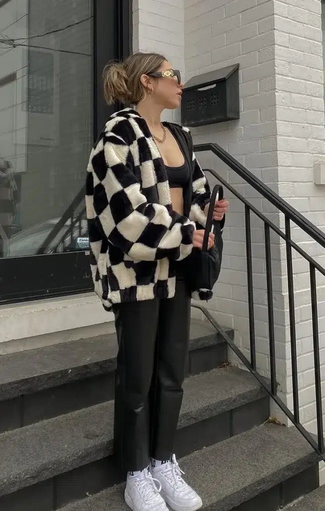 A petite woman standing on city steps outside a white brick building, wearing an oversized black and white large-check fluffy teddy jacket worn open over a black fitted crop sports bra, paired with sleek black faux leather straight-leg pants and white sneakers, with a gold chain necklace and oversized square sunglasses, holding a black bag, with an urban street visible in the background.