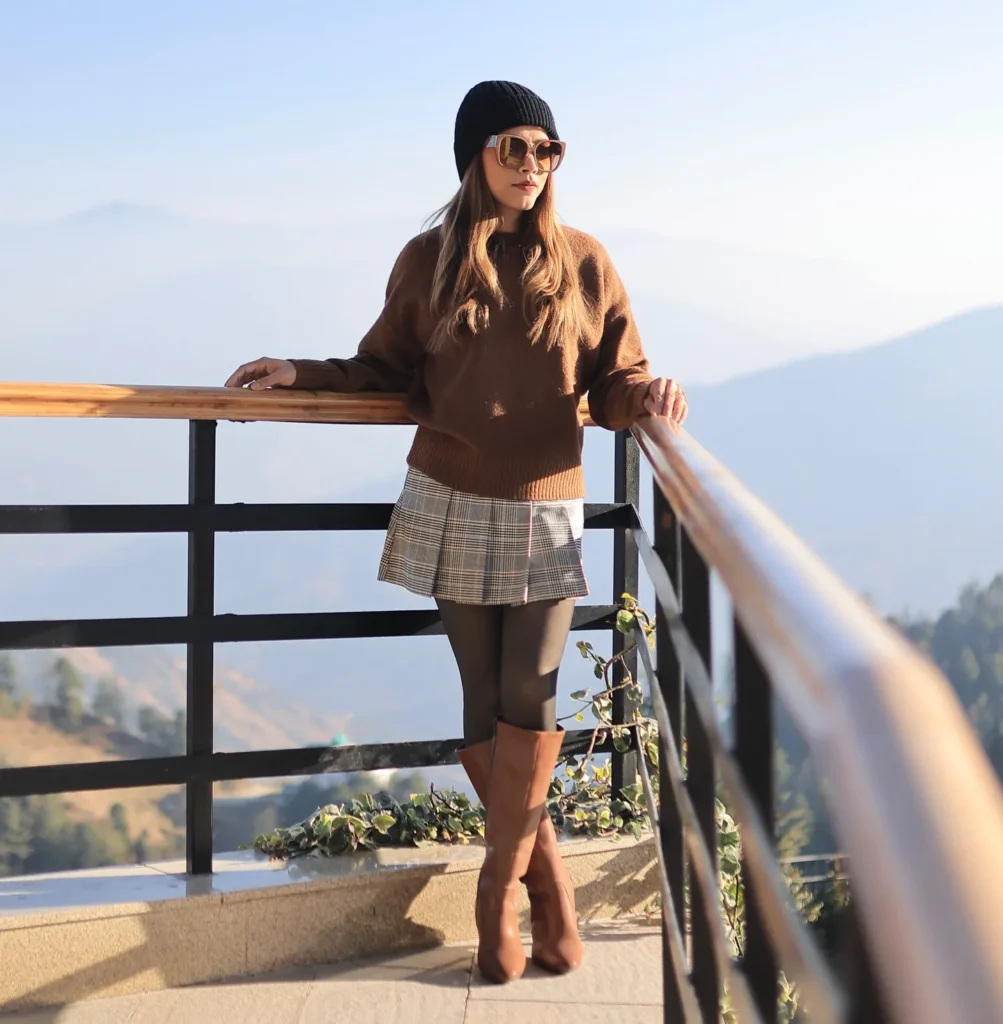 Petite woman with long light brown hair leaning against an iron railing on a sunlit mountain terrace with hazy blue mountain peaks in the background, wearing a boxy chocolate brown oversized knit sweater, a grey plaid pleated mini skirt, dark charcoal opaque tights, tan camel knee-high heeled boots, a black ribbed beanie pushed slightly back on the head, and large oversized brown-tinted sunglasses.