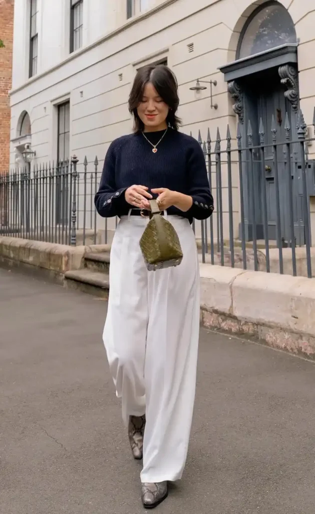 A petite woman walking on a quiet European cobblestone street, wearing a navy chunky ribbed knit sweater with gold button cuff details and a dark belt at the waist, paired with high-waisted white wide-leg fluid trousers and snakeskin-print ankle boots, holding a small olive green woven bag, with a cream Georgian building and black iron fence in the background.