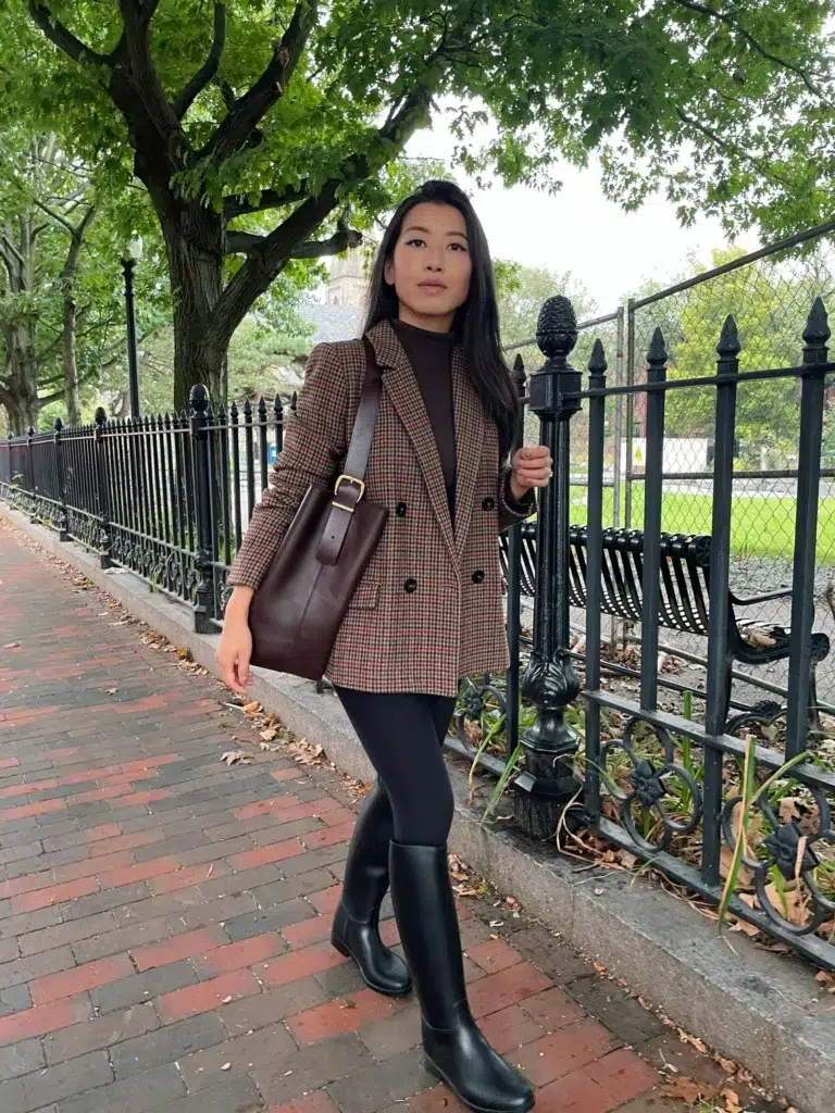 Petite woman in a warm brown houndstooth double-breasted blazer over a dark chocolate turtleneck, paired with black leggings, tall flat black boots, and a deep burgundy structured leather tote with a gold buckle, standing on a brick city sidewalk beside an iron fence with autumn trees in the background.