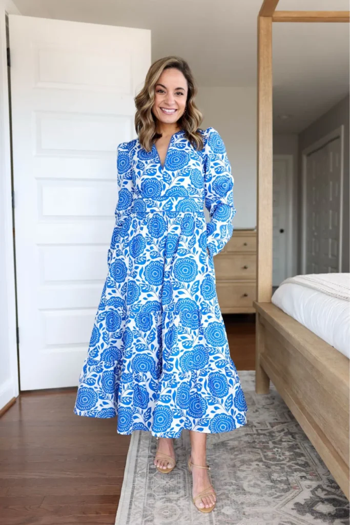 Smiling woman standing in a bright bedroom interior in a bold cobalt blue and white large-scale block floral print long-sleeve V-neck tiered midi dress hitting at mid-calf, paired with nude tan strappy low-heeled sandals, standing on a patterned area rug with a wooden bed frame and white door visible in the background.