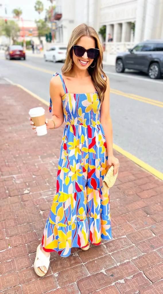 A petite woman wearing a cobalt blue maxi dress with a bold large-scale tropical floral print in yellow, red, pink, and white, featuring a smocked bodice, tie-bow shoulder straps, and a graduated tiered skirt with increasingly large motifs toward the hem, styled with white cross-strap platform espadrille mule sandals, a small natural woven half-moon clutch, large gold hoop earrings, and a gold bracelet, photographed walking on a brick sidewalk in an urban street setting.