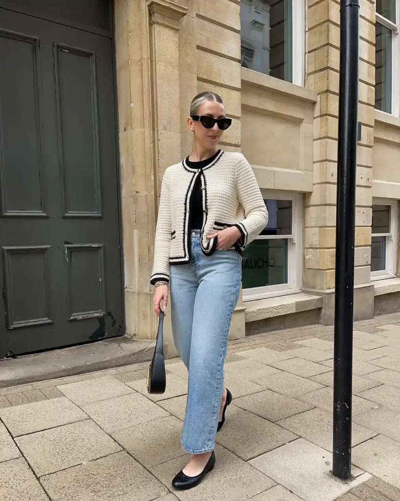 A woman standing on a stone-paved city street beside a dark grey door and sandstone building, wearing a cream textured open-knit cardigan with black contrast trim along the collar, front edges, cuffs, and pockets, layered over a fitted black top, paired with light-wash ankle-length straight-leg jeans and black ballet flats, carrying a small black structured top-handle bag and wearing dark cat-eye sunglasses with hair slicked back.
