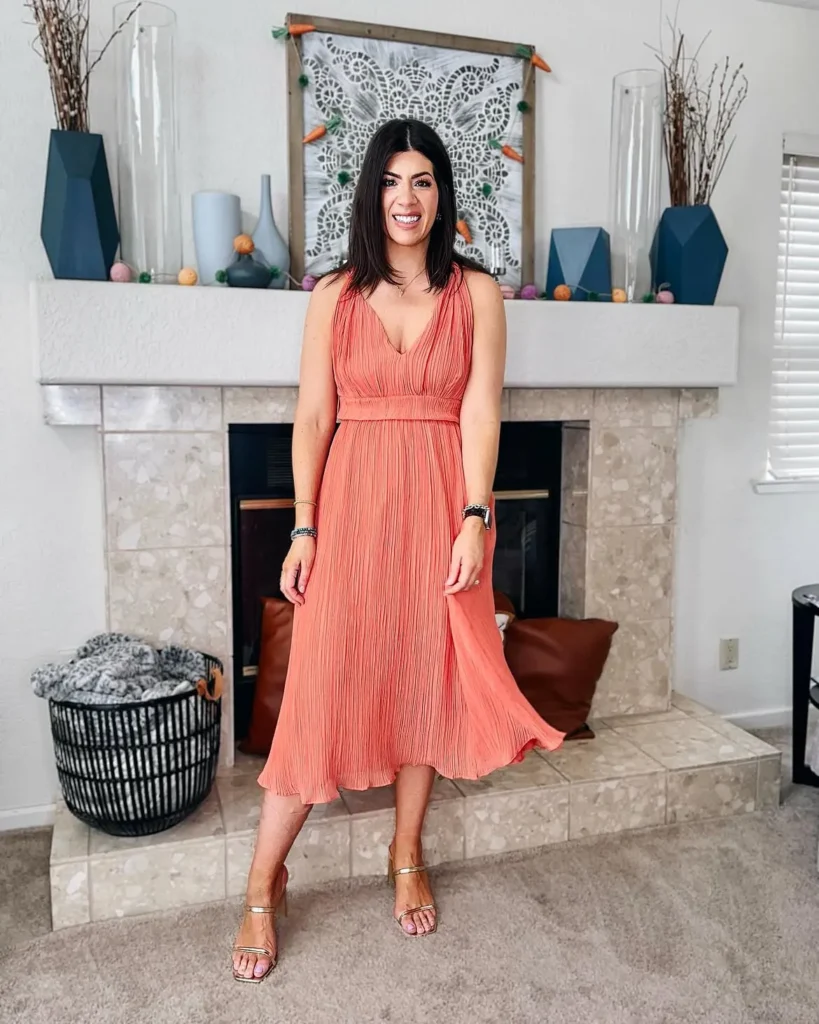 Petite woman in a coral salmon-toned pleated empire waist midi dress with a V-neckline, delicate shoulder tie straps, and a banded waist detail, wearing gold thin-strapped heeled sandals and stacked bracelets, smiling while standing in front of a tiled fireplace mantel inside a bright living room.