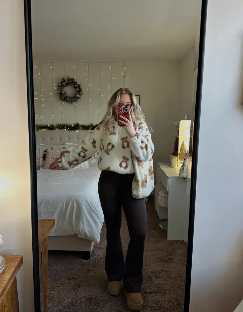 Petite woman with wavy blonde hair taking a mirror selfie in a cozy Christmas-decorated bedroom with fairy lights, a holiday wreath, and garland on the headboard, wearing an oversized cream fluffy sherpa zip-up sweatshirt with an allover brown teddy bear print, fitted black flare leggings, and tan platform clog-style shoes, holding a phone with a red polka dot case.