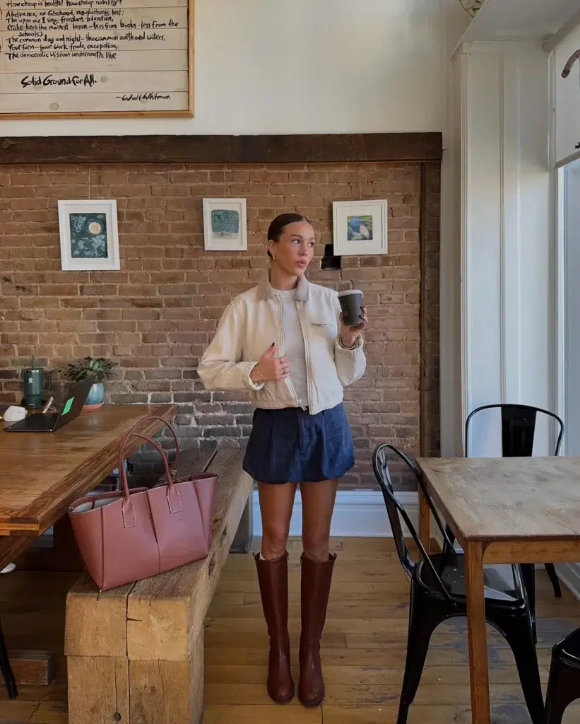 Petite woman in a cream ivory zip-front bomber jacket with a ribbed hem and small chest embroidery detail, worn over a dark indigo denim mini skirt with a soft relaxed fit, paired with tall cognac warm brown leather knee-high riding boots with a flat heel, bare-legged between the mini hem and the boot shaft, standing inside a cozy café with exposed brick walls, wooden communal tables, and small framed artworks on the wall, with a dusty rose structured leather tote bag placed on the wooden bench beside her, holding a dark paper coffee cup, hair pulled back in a sleek low bun with gold hoop earrings.