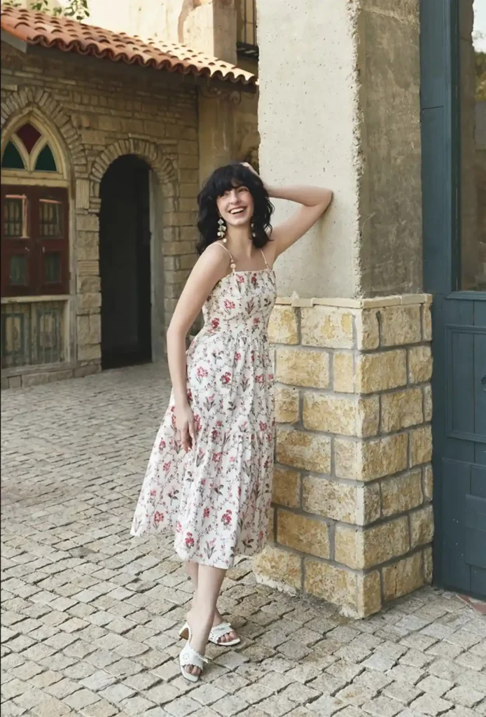 Petite woman with short curly dark hair and a blunt fringe in a cream off-white midi dress covered in small scattered red and pink botanical sprig florals, featuring thin spaghetti straps, a fitted corset-style boned bodice, and a full tiered midi skirt, wearing white heeled mule sandals with decorative cutout detail and large tiered drop earrings in warm cream and gold tones, laughing and leaning against a sandy stone pillar on a cobblestone street with Mediterranean stone arch architecture in the background.
