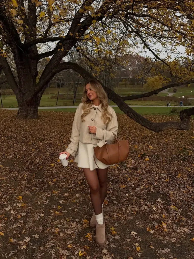 Petite woman in a full cream monochrome outfit consisting of an oversized cropped boxy wool-look button-front jacket with a flat collar, worn over an ivory flared mini skirt, paired with sheer nude tights, white chunky ribbed crew socks at the ankle, and tan camel suede low-heeled ankle boots, carrying a large structured cognac tan leather open-top tote bag, holding a white takeaway coffee cup, standing on a leaf-covered path in a park with golden autumn trees visible in the background on an overcast fall day.