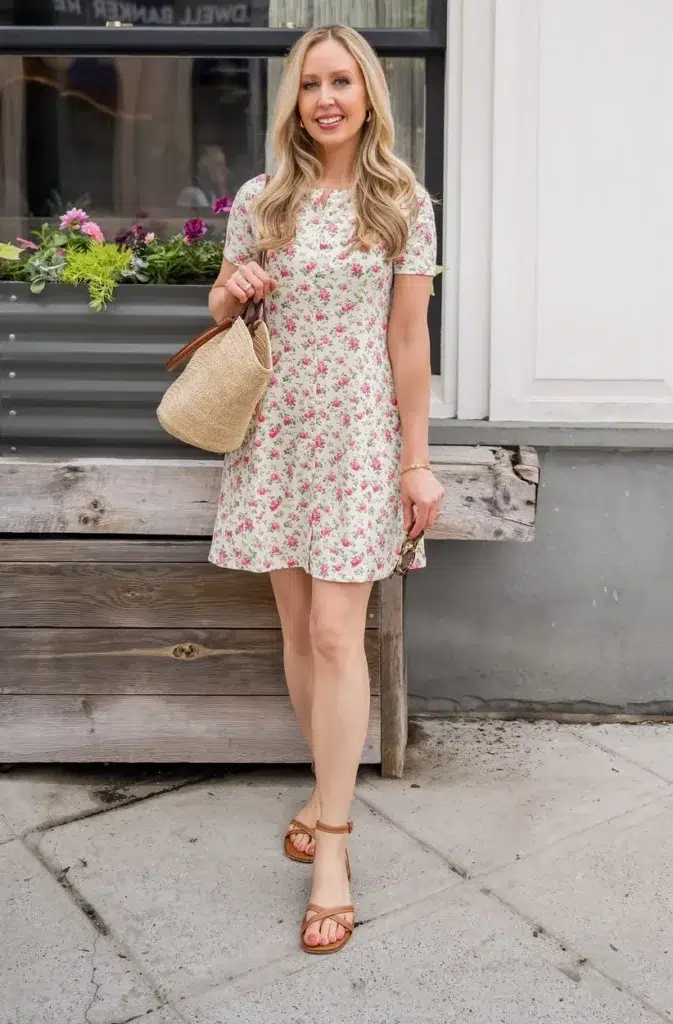 Petite woman with long blonde wavy hair in a cream off-white short-sleeve A-line mini dress with a small scattered pink ditsy rose floral print, short puff sleeves, and a rounded neckline, wearing tan strappy block heel ankle strap sandals and carrying a small round woven straw basket bag with tan leather handles and wrist strap, with a gold bracelet and small gold stud earrings, standing on a city sidewalk in front of a café storefront with window boxes of purple and pink flowers in the background.