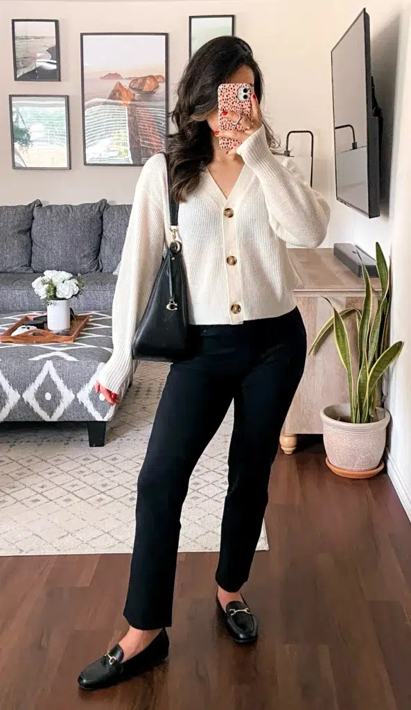 Petite woman with long dark wavy hair taking a mirror selfie in a bright living room with a grey sectional sofa, geometric area rug, and gallery wall of landscape prints, wearing a cream chunky ribbed V-neck cardigan fully buttoned with large gold button detail, black slim straight-leg cropped ankle pants, black horsebit flat loafers with gold hardware, and a black shoulder hobo bag with gold clasp, with a snake plant visible in the background.
