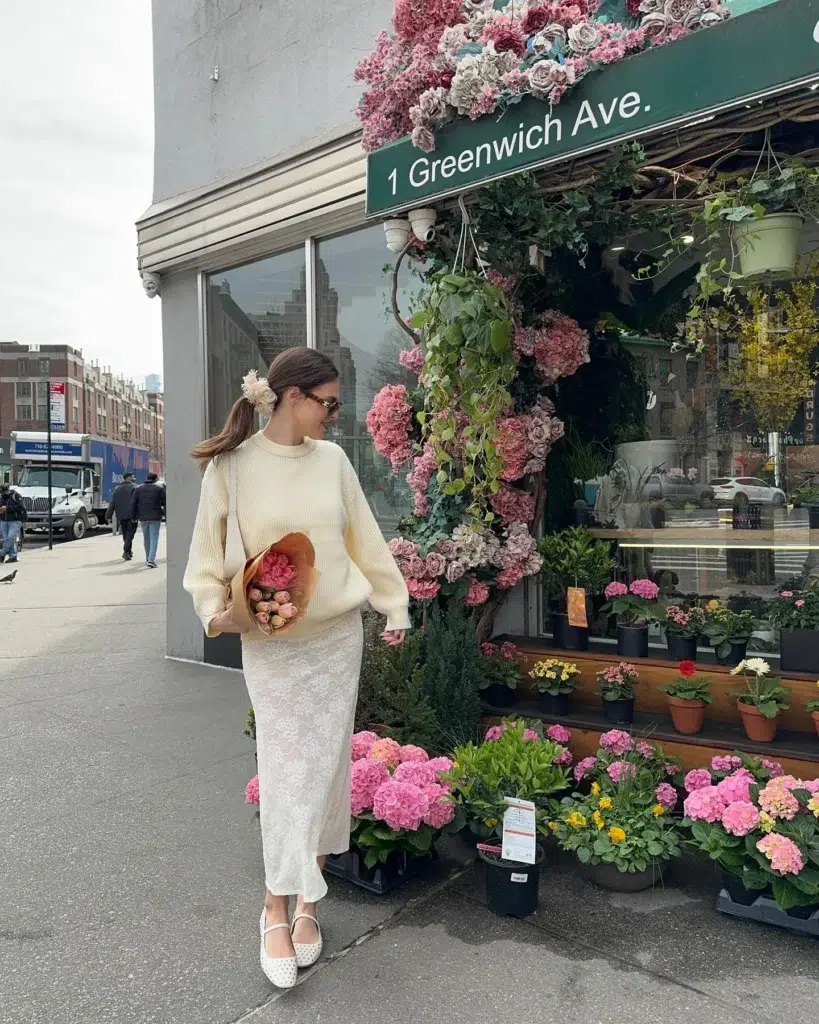 Petite woman walking past a corner flower shop, wearing a cream ribbed knit sweater over a matching ivory lace maxi skirt, white polka-dot ballet flats, a white shoulder bag, and a floral hair scrunchie, holding a bouquet of pink tulips, with hydrangeas and roses around her.