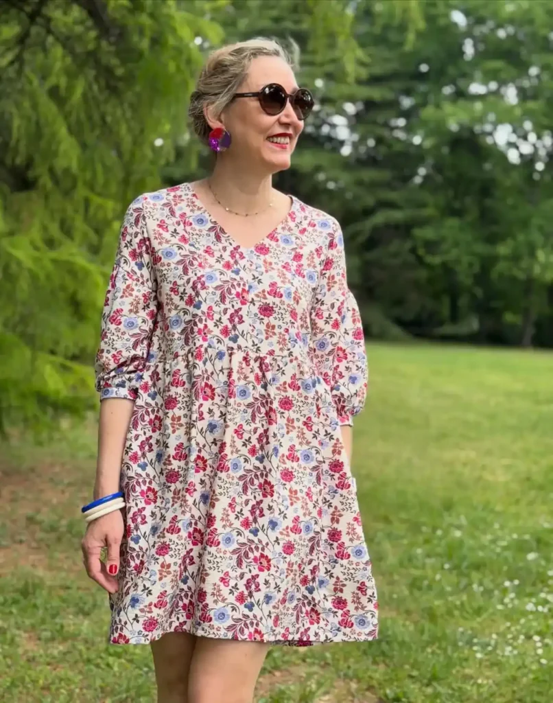 Woman in a cream babydoll mini dress covered in a multi-toned floral print in pink, lavender, burgundy, and rust, with a V-neckline and gathered bodice, wearing large round purple and red statement earrings, tortoiseshell round sunglasses, a delicate gold chain necklace, and blue and white stacked bangle bracelets, smiling outdoors in a lush green garden setting.