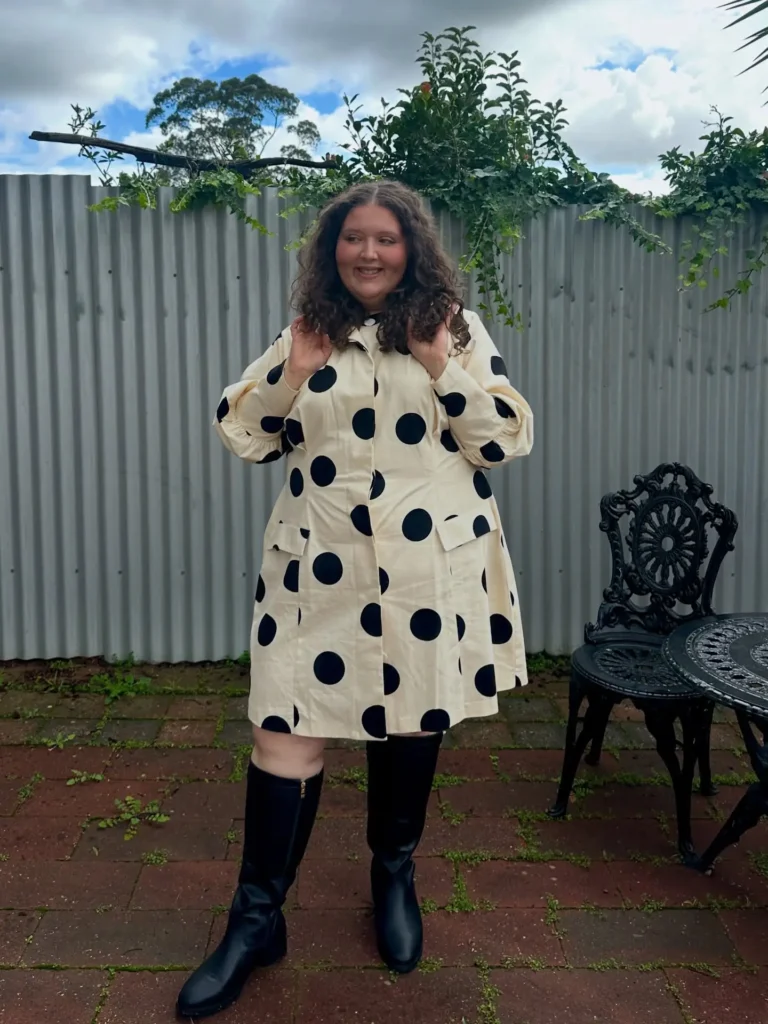 Curvy petite woman standing on a brick patio outdoors wearing a cream oversized A-line polka dot shirt dress with large black dots, a button-front placket, collar, and long gathered puff sleeves, paired with tall black knee-high boots with a small heel and side zipper, with natural curly brown hair worn loose, in front of a corrugated metal fence with trailing vines.