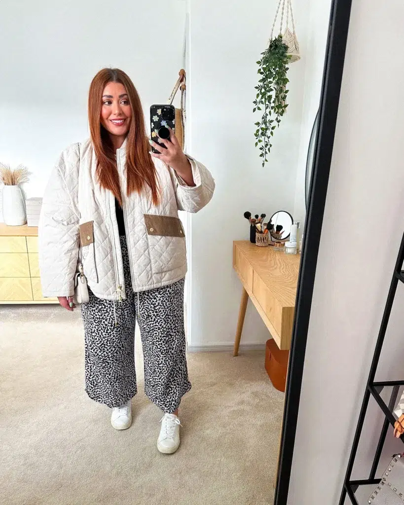 Woman with long straight auburn hair taking a mirror selfie in a bright modern bedroom with a wooden vanity and hanging trailing plant in a macramé hanger, wearing a cream diamond-quilted collarless zip-front jacket with tan suede patch pockets and gold hardware, black and white small ditsy print wide-leg culottes, white clean leather lace-up sneakers, and a small white shoulder bag, smiling at the camera.