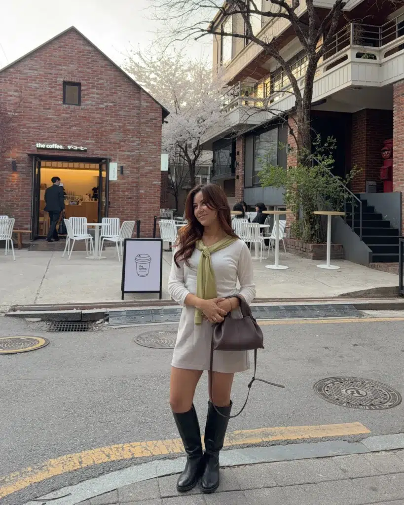 Petite woman standing on a city street near a brick café, wearing a cream long-sleeve sweater dress with an olive-green sweater knotted over the shoulders, tall black riding boots, and holding a chocolate-brown top-handle bag, cherry blossoms behind her.