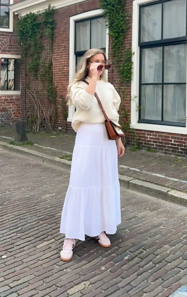 Petite woman walking on a cobblestone street, wearing a cream sweater loosely tucked into a white tiered maxi skirt, pale pink gum-sole sneakers, a tan leather crossbody bag, and sunglasses, with an ivy-covered brick building behind her.