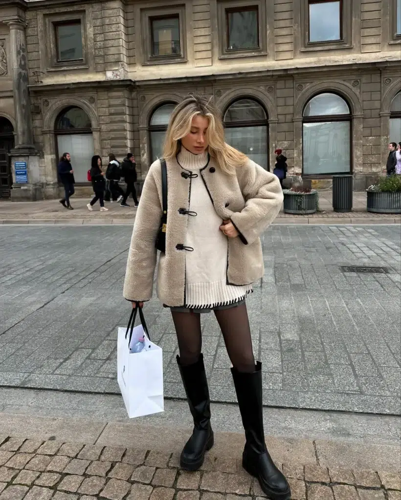 Petite woman with blonde hair standing on a cobblestone European city square in front of a grand stone building with arched windows, wearing an oversized cream faux-shearling teddy jacket with black toggle closures and black contrast edge trim, layered over a cream chunky ribbed turtleneck sweater with the ribbed hem visible below the jacket, a grey micro mini skirt, dark sheer tights, and tall chunky-soled black platform knee-high boots, with a black crossbody bag on her shoulder and a white shopping bag in hand.