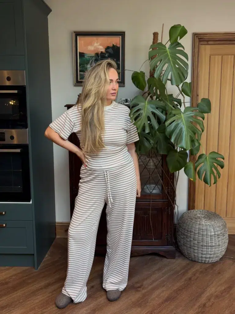 Petite Woman with long blonde wavy hair standing in a stylish home with dark green kitchen cabinetry and a large monstera plant, wearing a matching cream and taupe fine horizontal-stripe jersey co-ord set with a fitted short-sleeve cropped crew-neck top and wide-leg drawstring-waist trousers with a floor-grazing hem, with short taupe ankle boots just visible below the wide trouser hem.