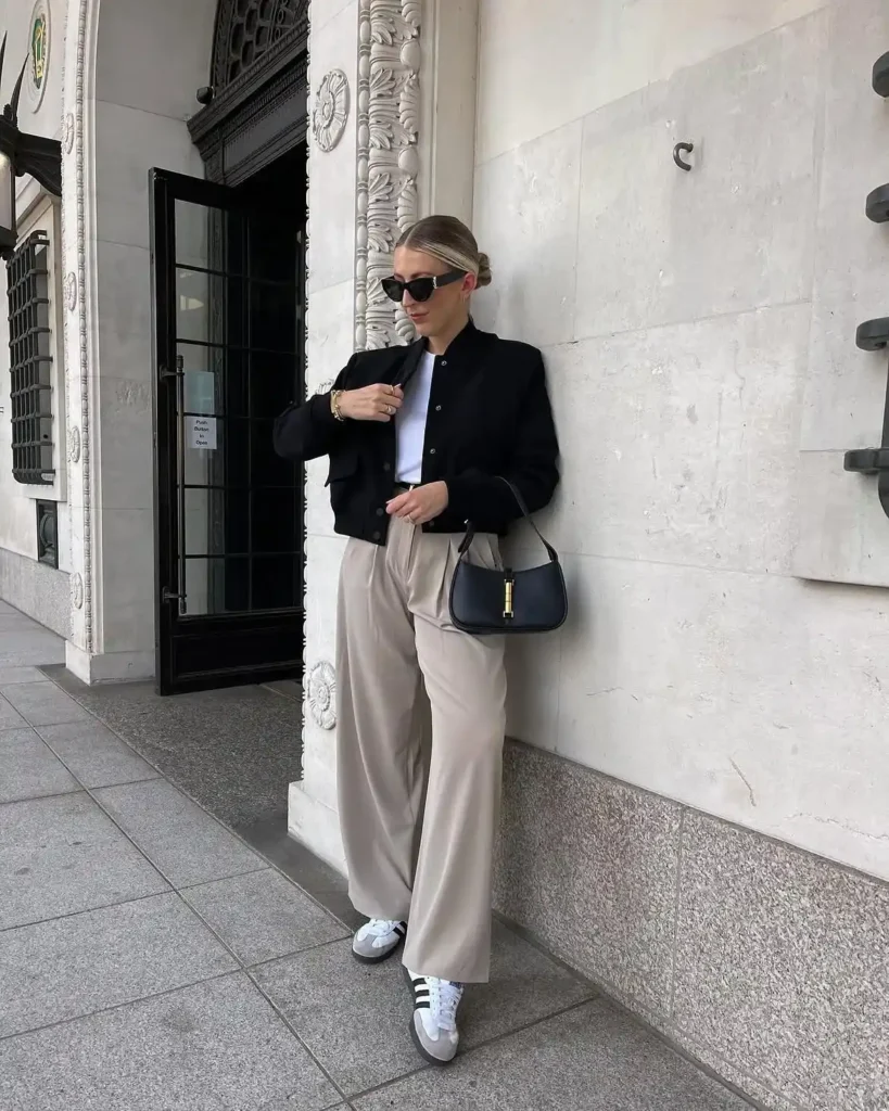 A woman leaning against an ornate cream stone building facade on a city pavement, wearing a fitted black cropped bomber jacket with snap button closure over a white fitted tee tucked into high-waisted wide-leg tailored trousers in a stone beige tone, with white and black stripe low-profile trainers and a small black structured shoulder bag with gold hardware detail, wearing dark cat-eye sunglasses with hair in a low bun.