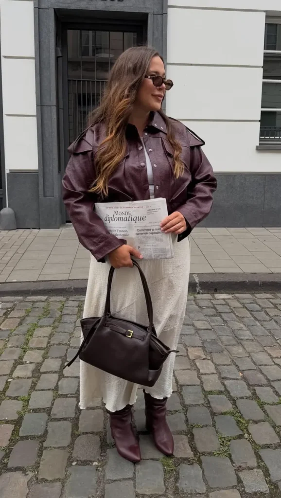 A petite woman standing on a cobblestone European street outside a white and grey building, wearing a deep oxblood cropped leather jacket with wide lapels over a dark button-front shirt, paired with a white cream crinkled textured midi skirt falling to mid-calf, with dark burgundy ankle boots and a structured dark chocolate leather tote bag, holding a French newspaper and wearing small oval sunglasses.