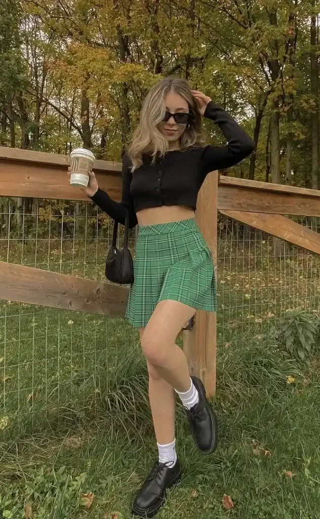 A petite woman leaning against a wooden post fence in an autumn outdoor setting with trees showing orange and yellow foliage, wearing a fitted black long-sleeve button-front crop cardigan top paired with a green and black tartan pleated mini skirt, white crew socks, and chunky black lace-up platform boots, holding a takeaway coffee cup with a small black shoulder bag at her side and dark rectangular sunglasses.