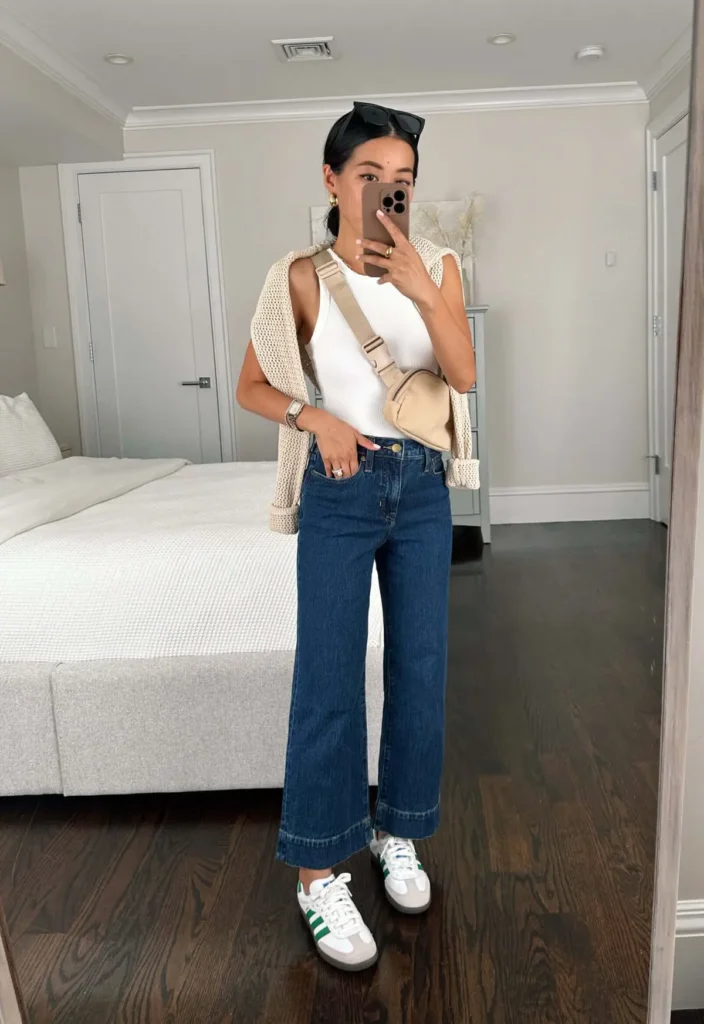 Mirror selfie of a petite woman in cropped medium-wash wide leg jeans, a white ribbed tank, a cream open-knit vest, a sand mini belt bag worn crossbody, and white and green retro sneakers
