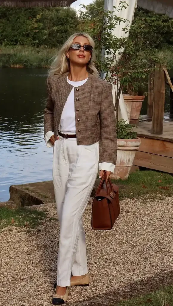 A woman walking on a gravel path beside a still lake, wearing a cropped brown houndstooth collarless jacket with gold button front over a white fitted crew-neck tee, paired with high-waisted relaxed wide-leg white linen trousers and a slim cognac leather belt at the waist, carrying a structured cognac brown leather tote bag and wearing narrow oval sunglasses, with green trees and a wooden dock visible in the background.