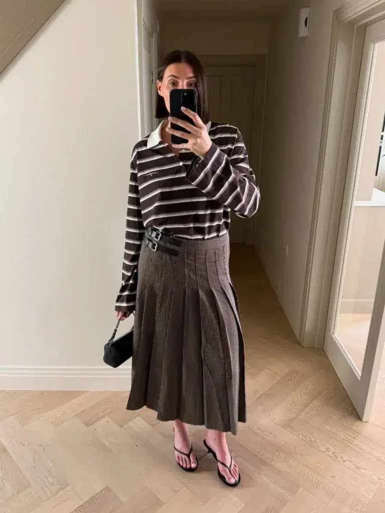 Petite woman in an oversized dark chocolate and white horizontal-striped rugby polo shirt with a white collar, worn bloused over a brown micro-check pleated midi skirt and cinched at the waist with a black leather belt with a bold buckle, paired with black strappy low-heeled thong sandals and a small black mini bag held by a thin strap, taking a mirror selfie in a bright home hallway with warm wood herringbone flooring and neutral walls.