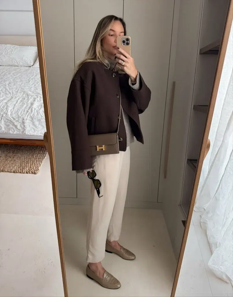Petite woman in a dark chocolate brown boxy collarless wool-look jacket with large snap button closures and front patch pockets, layered over a warm gray ribbed knit sweater visible at the neckline and cuffs, paired with slim tapered cream ivory trousers falling cleanly to the ankle, wearing flat taupe greige quilted loafers, with a structured tan taupe crossbody bag featuring a large gold H-shaped clasp worn across the body, holding dark tortoiseshell sunglasses in one hand, taking a mirror selfie in a minimalist bedroom with soft gray wardrobe doors and a gold-framed full-length mirror.