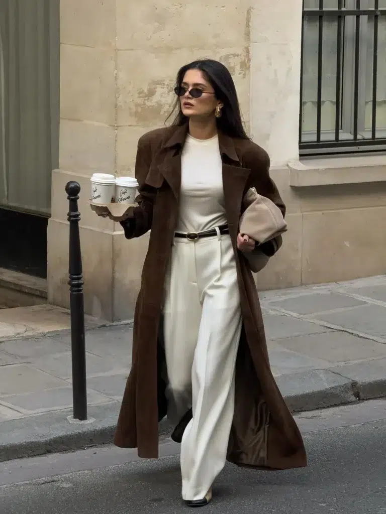 Petite woman with long straight dark hair walking on a narrow Parisian stone street past a classic Haussmann building and an iron bollard, wearing a rich dark chocolate brown longline open suede-look coat with wide lapels hanging fully open as two side panels, a fitted cream ivory high-neck base layer top, floor-length cream wide-leg tailored trousers with a center crease, a slim black leather belt with a small gold oval buckle at the natural waist, small narrow dark oval sunglasses, sculptural gold earrings, a soft blush nude pouchy bag tucked under one arm, holding two branded coffee cups in a cardboard carrier tray.