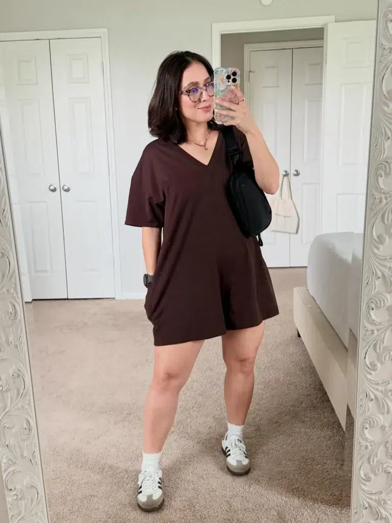Curvy petite woman taking a bedroom mirror selfie wearing a deep chocolate brown oversized V-neck short-sleeve romper with side pockets, paired with white ankle socks and white and black Adidas sneakers, carrying a black crossbody sling bag worn diagonally across the body, styled with tortoiseshell glasses, layered gold necklaces, a black smartwatch, and a short dark bob hairstyle.