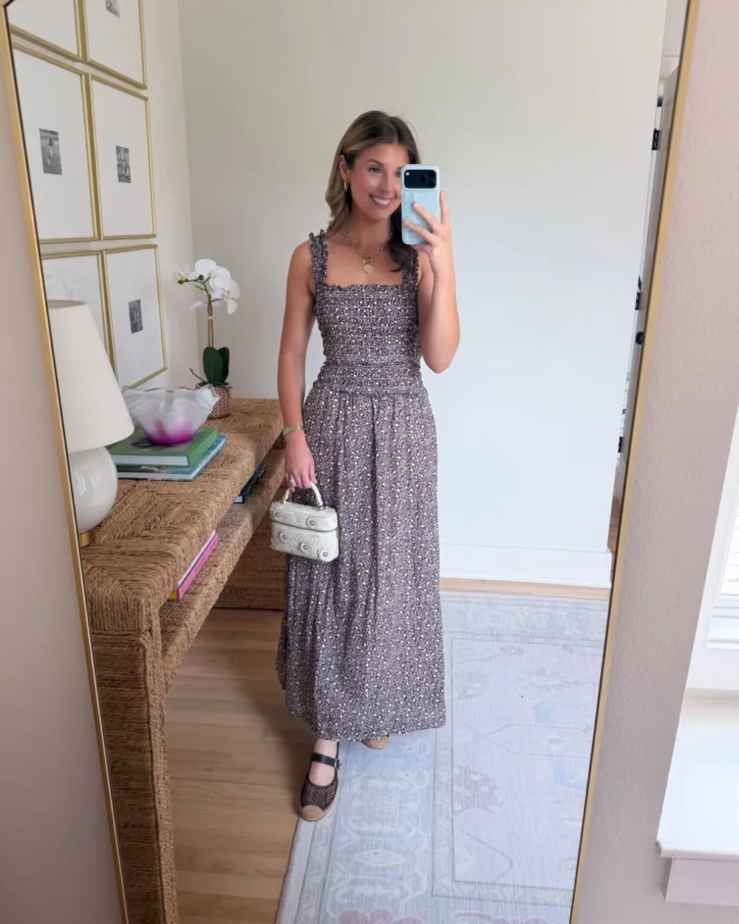 Petite woman taking a mirror selfie in a dark plum-brown ditsy floral smocked maxi dress with ruffle straps and a square neckline, wearing dark espadrille wedge Mary Jane sandals and holding a small cream embellished structured box bag, with a layered gold coin pendant necklace, reflected in a gold-framed floor mirror with a rattan console table and white orchid visible in the background.